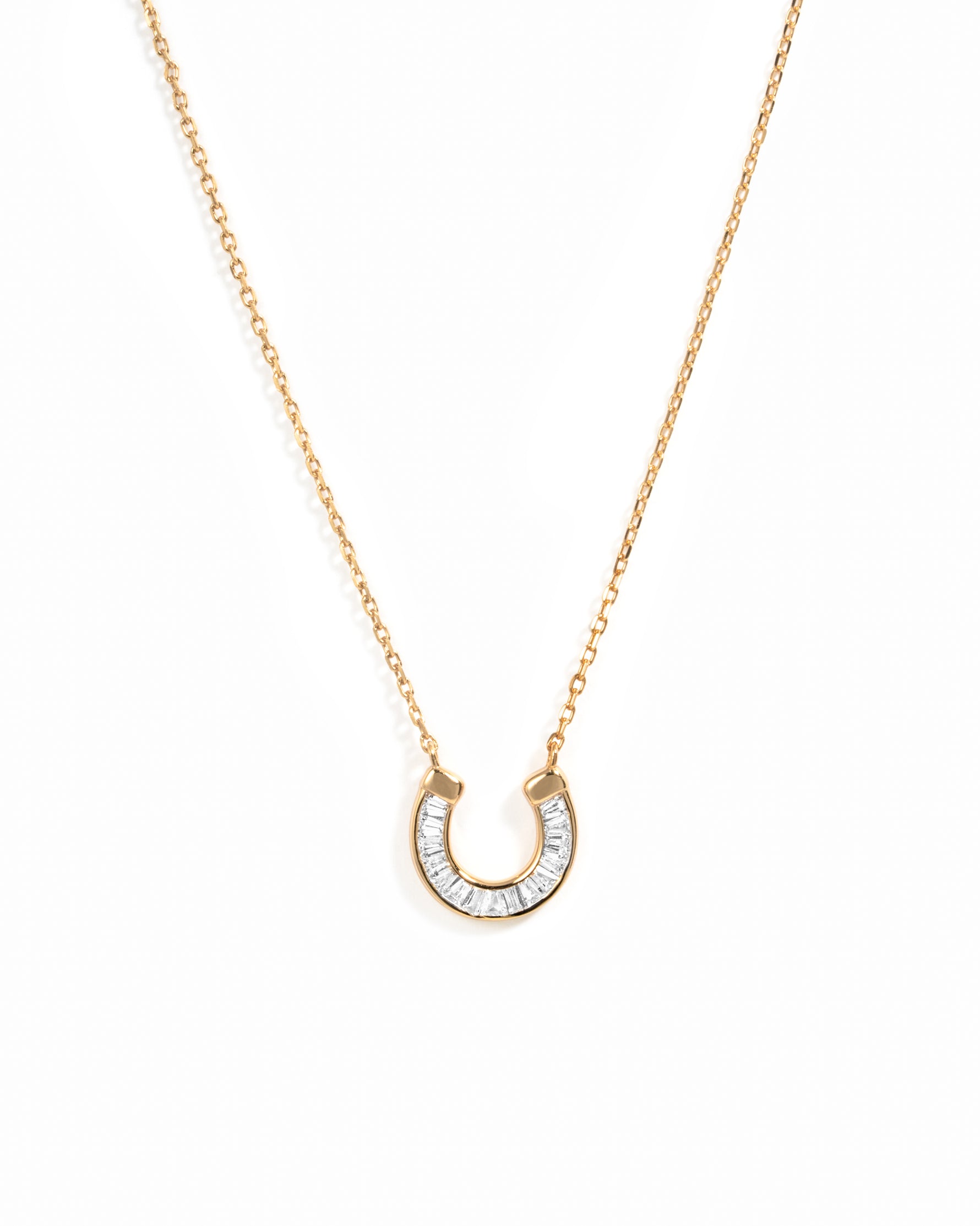 A delicate gold chain necklace featuring a horseshoe-shaped pendant paved with baguette cut stones, set against a plain white background.