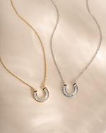Two delicate horseshoe-shaped necklaces with pavé-set baguette cz stones, one in yellow gold and one in silver, displayed on a soft beige satin fabric background.