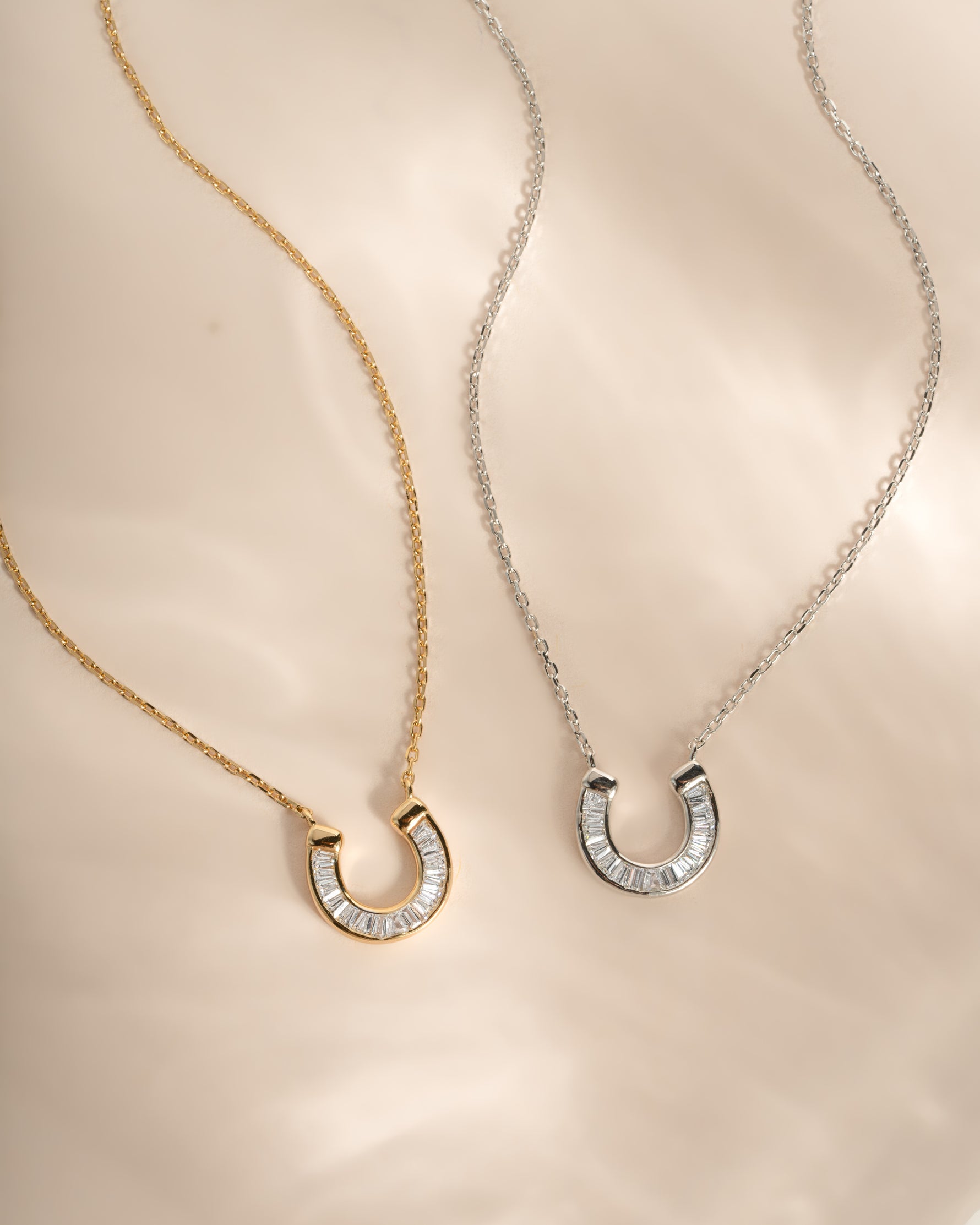 Two delicate horseshoe-shaped necklaces with pavé-set baguette cz stones, one in yellow gold and one in silver, displayed on a soft beige satin fabric background.