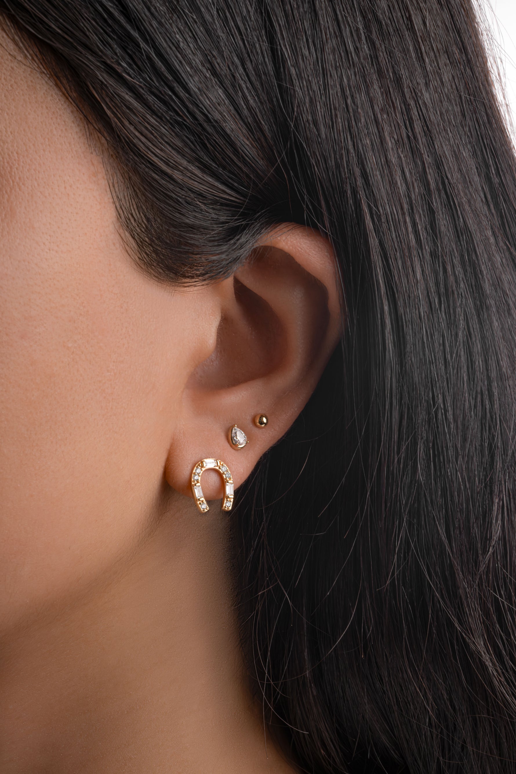 A close-up of a model's ear showcasing a gold horseshoe shaped hoop earring adorned with baguette and round stones
