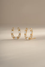 A pair of gold horseshoe shaped stud earrings embellished with clear baguette and round diamonds, displayed on a neutral beige background.