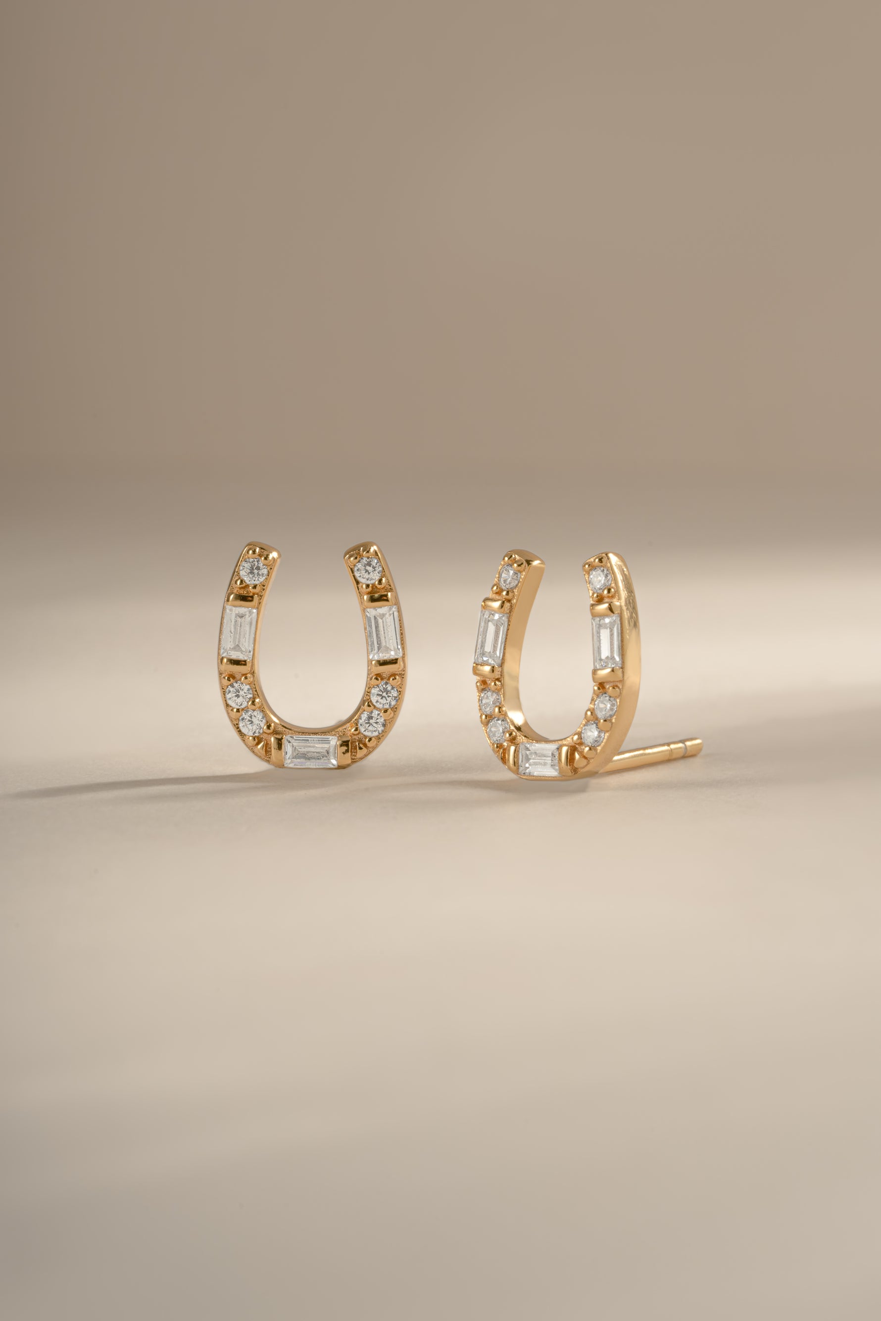 A pair of gold horseshoe shaped stud earrings embellished with clear baguette and round diamonds, displayed on a neutral beige background.