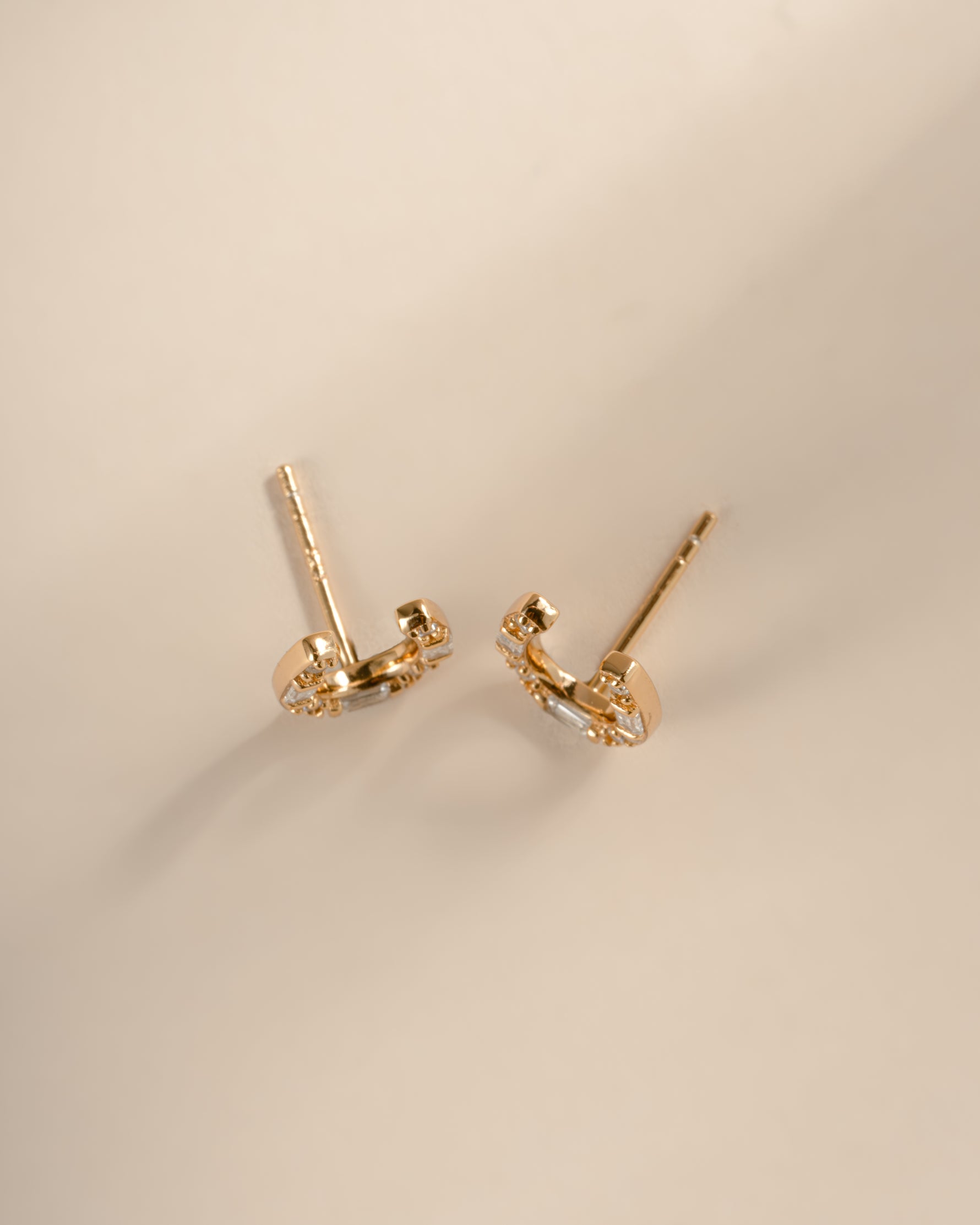 A top view of a pair of gold horseshoe shaped stud earrings embellished with clear baguette and round diamonds, displayed on a neutral beige background.