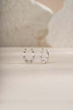 A pair of silver horseshoe shaped stud earrings embellished with clear baguette and round diamonds, displayed on a neutral beige background.