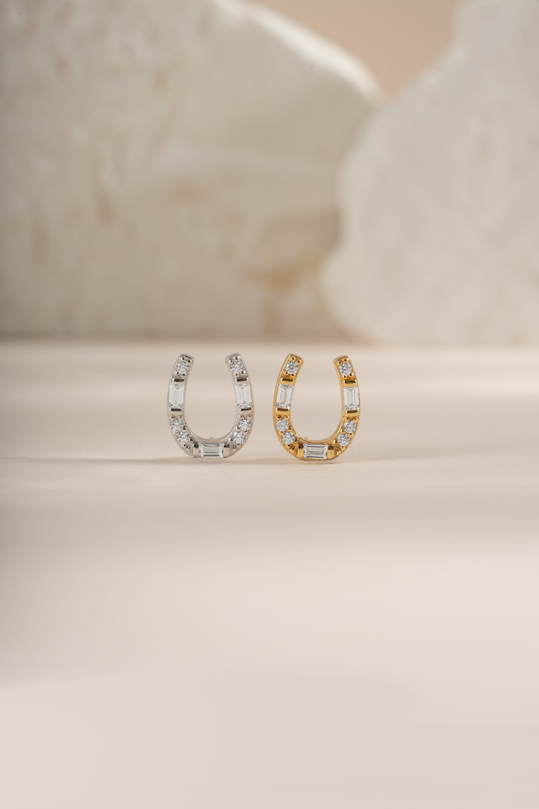 A gold and a silver horseshoe shaped stud earrings embellished with clear baguette and round diamonds, displayed on a neutral beige background.