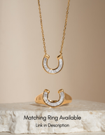Gold horseshoe pendant necklace with clear baguette stones, shown hanging and displayed on a stone slab against a warm beige background, with text noting a matching ring is available.