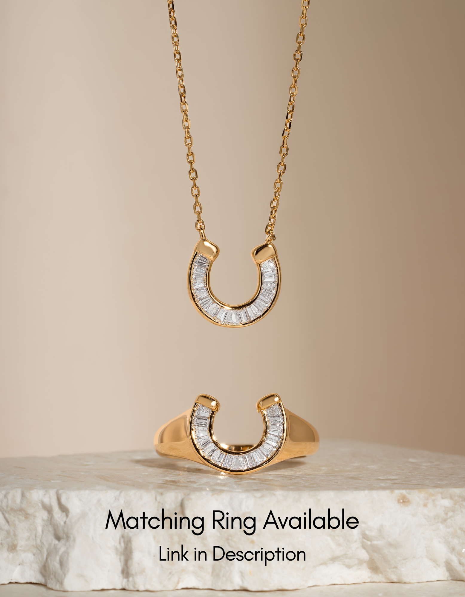 Gold horseshoe pendant necklace with clear baguette stones, shown hanging and displayed on a stone slab against a warm beige background, with text noting a matching ring is available.