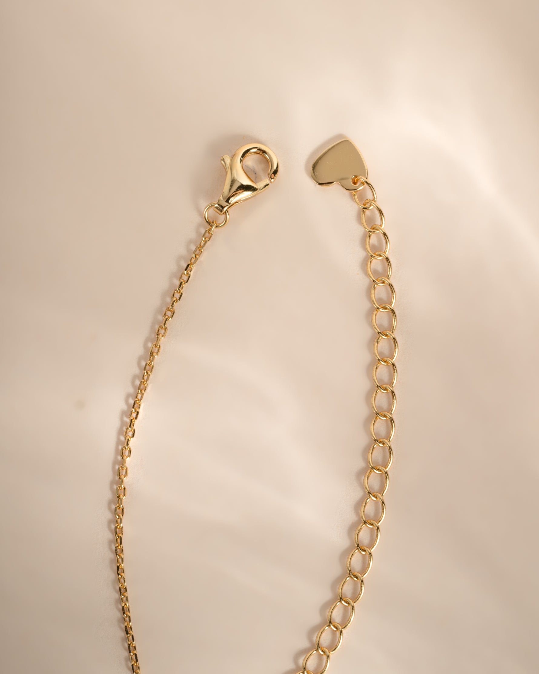 A gold-toned metal chain extender with a lobster clasp and a small heart-shaped tag, photographed on a soft beige background.