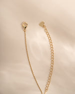A gold-toned chain extender strap with a lobster clasp and a small heart-shaped tag, displayed on a soft beige satin fabric background.
