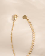 A gold-toned jewelry extension chain with a lobster clasp on one end and a small heart-shaped tag on the other, photographed on a soft cream background.