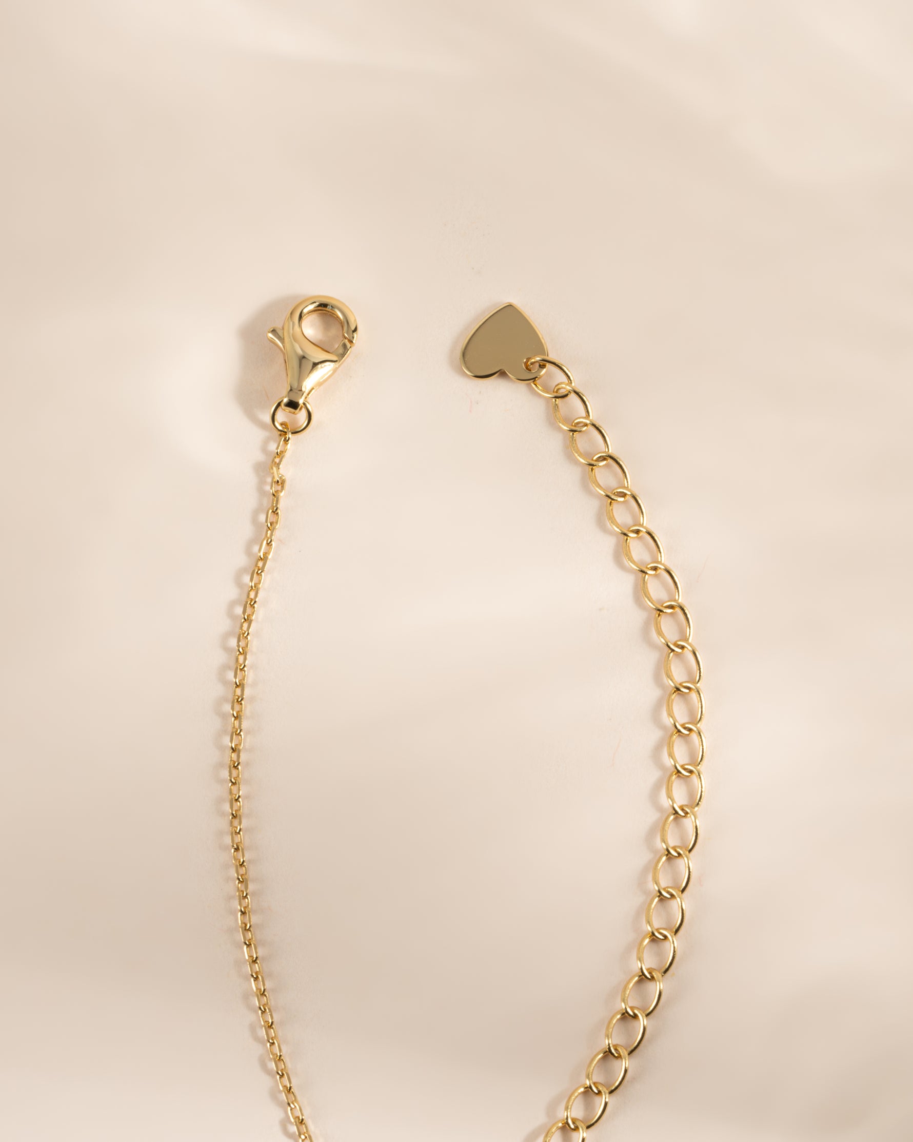A gold-toned jewelry extension chain with a lobster clasp on one end and a small heart-shaped tag on the other, photographed on a soft cream background.