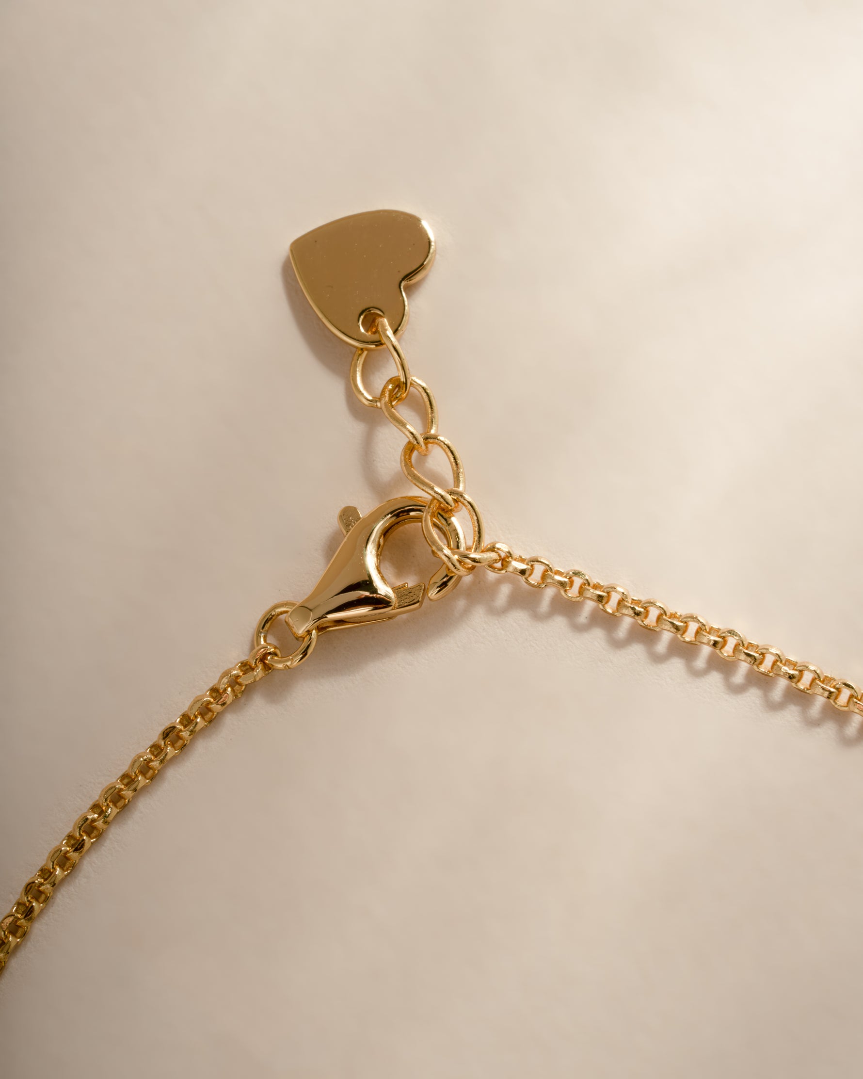 Close-up of a gold chain bracelet with a lobster clasp, an adjustable heart tag, and a small heart-shaped charm on a light beige background.
