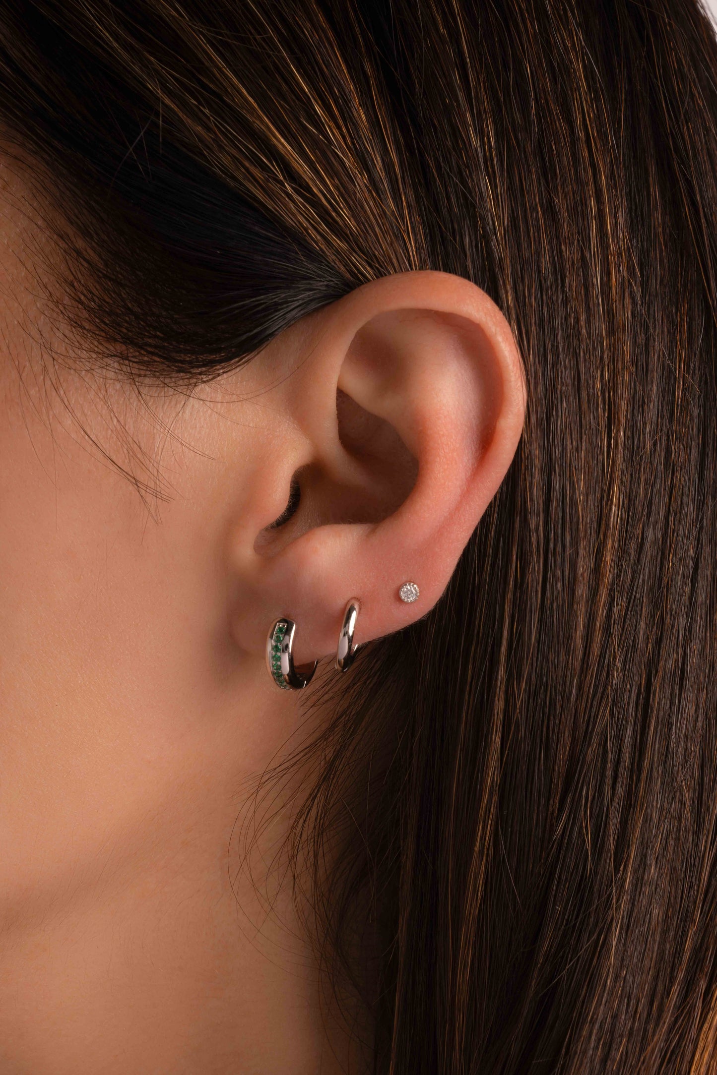 a close up of a model with three piercings, a small silver hoop earring with emerald gemstones in her first piercing, a small sterling silver huggie hoop in her second piercing, and a tiny diamond stud on her third piercing