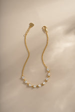 A delicate gold chain bracelet featuring a row of clear, tennis style pear stones set in gold, displayed on a soft beige background with warm, directional lighting.
