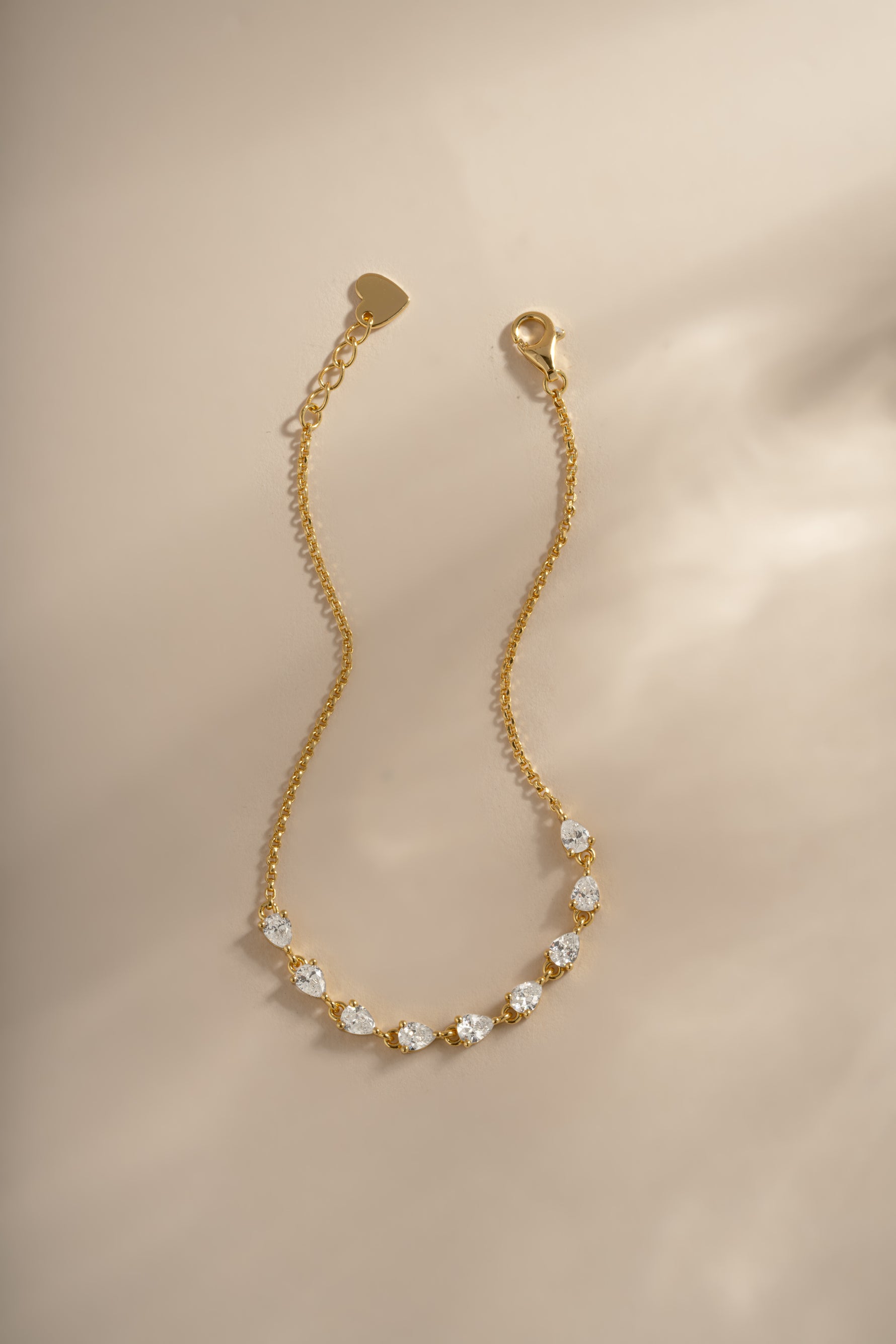 A delicate gold chain bracelet featuring a row of clear, tennis style pear stones set in gold, displayed on a soft beige background with warm, directional lighting.