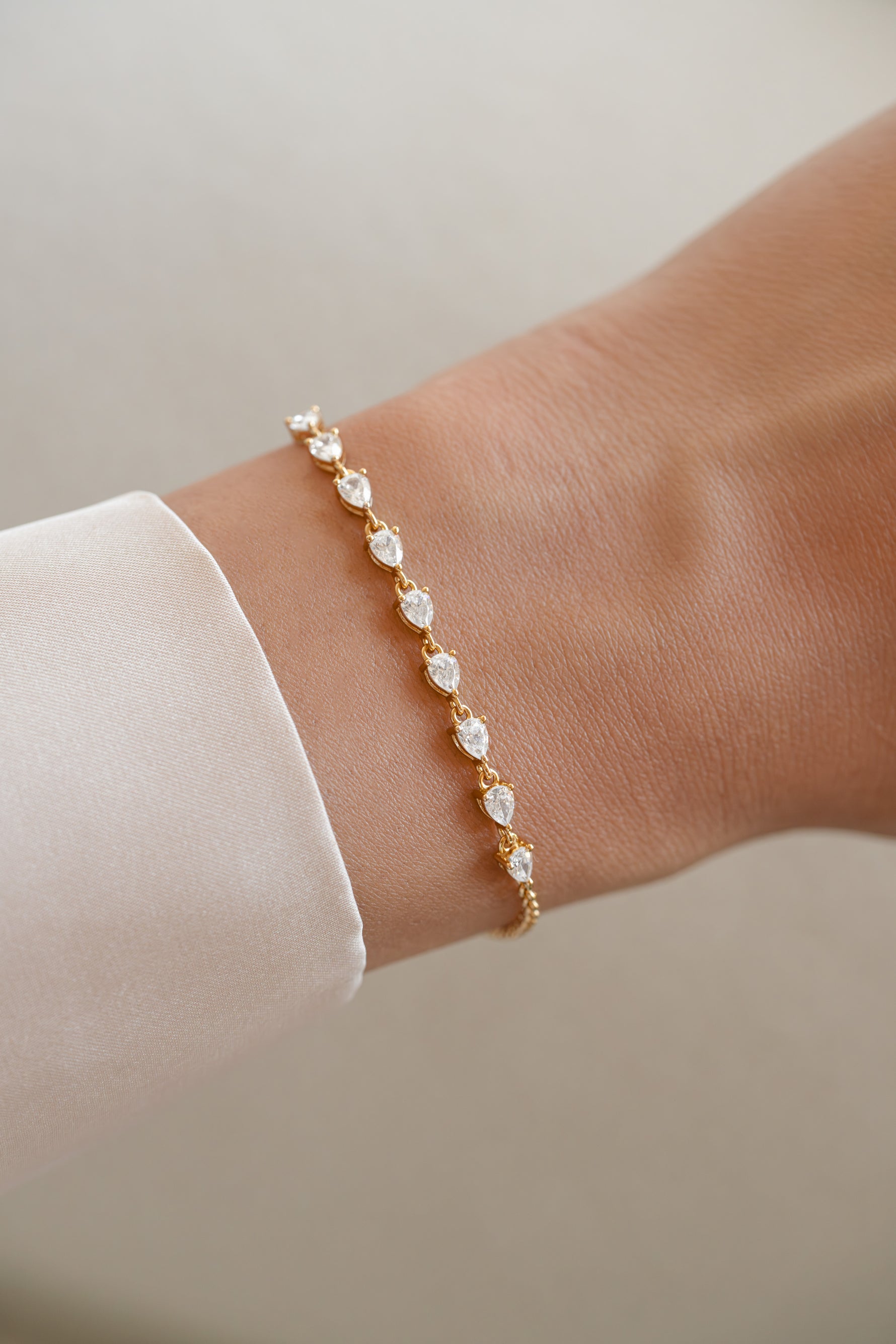 A delicate gold bracelet with small pear cut cz stones, worn on a person's wrist with a white sleeve visible.