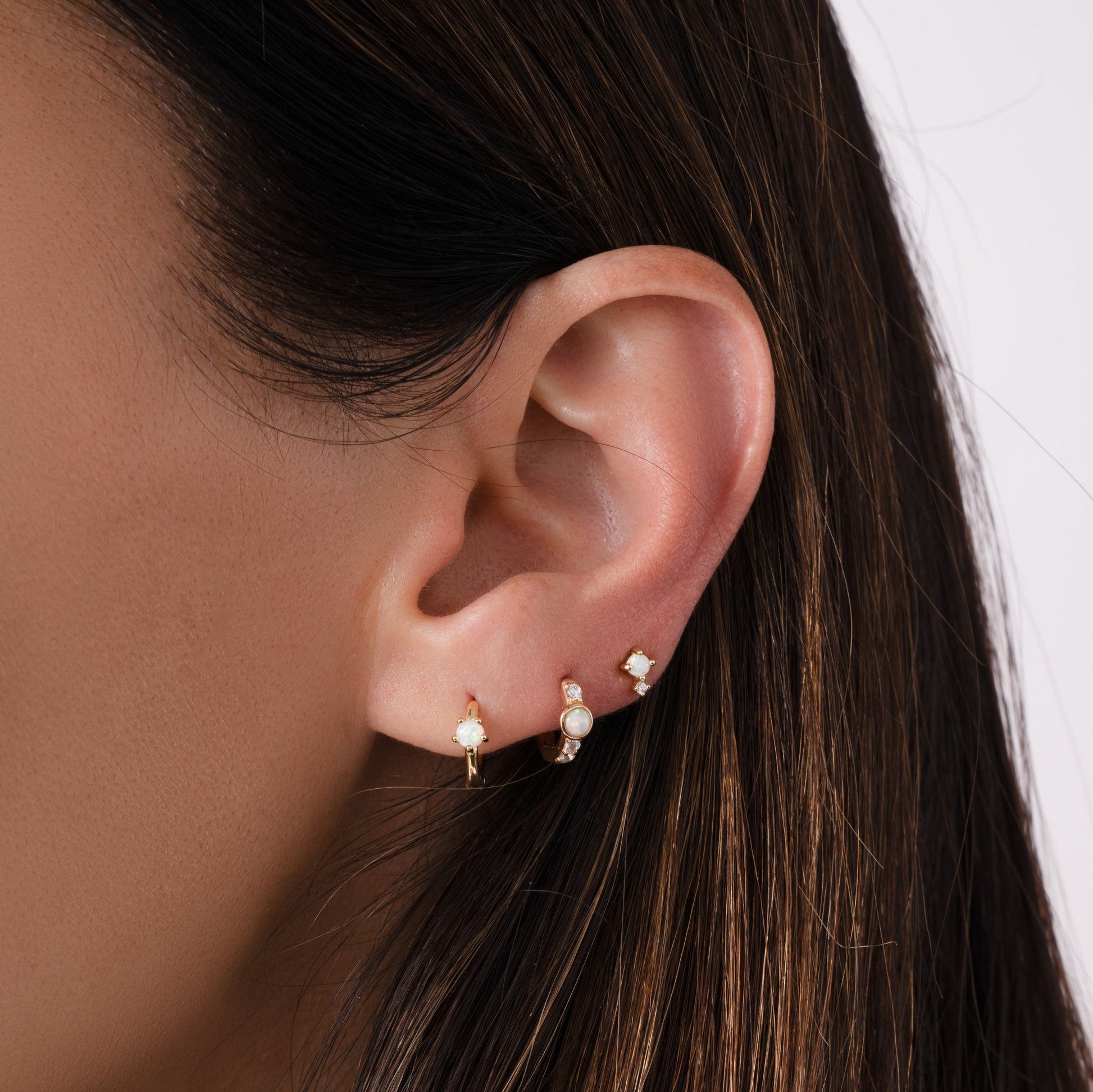 A closeup picture of a model wearing two small hoop earrings and one stud earrings. In the first piercing she is wearing small opal earrings, in the second is another small opal earring with diamonds, and in the third is a tiny opal stud earring. 