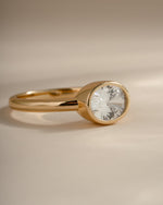 A gold ring with a bezel-set clear oval gemstone, photographed on a soft beige background.