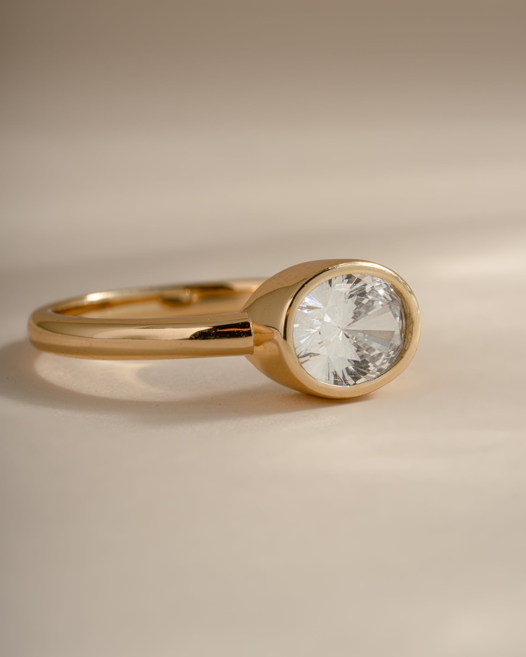 A gold ring with a bezel-set clear oval gemstone, photographed on a soft beige background.
