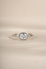 A silver ring with a bezel-set oval diamond gemstone, photographed on a soft beige background.