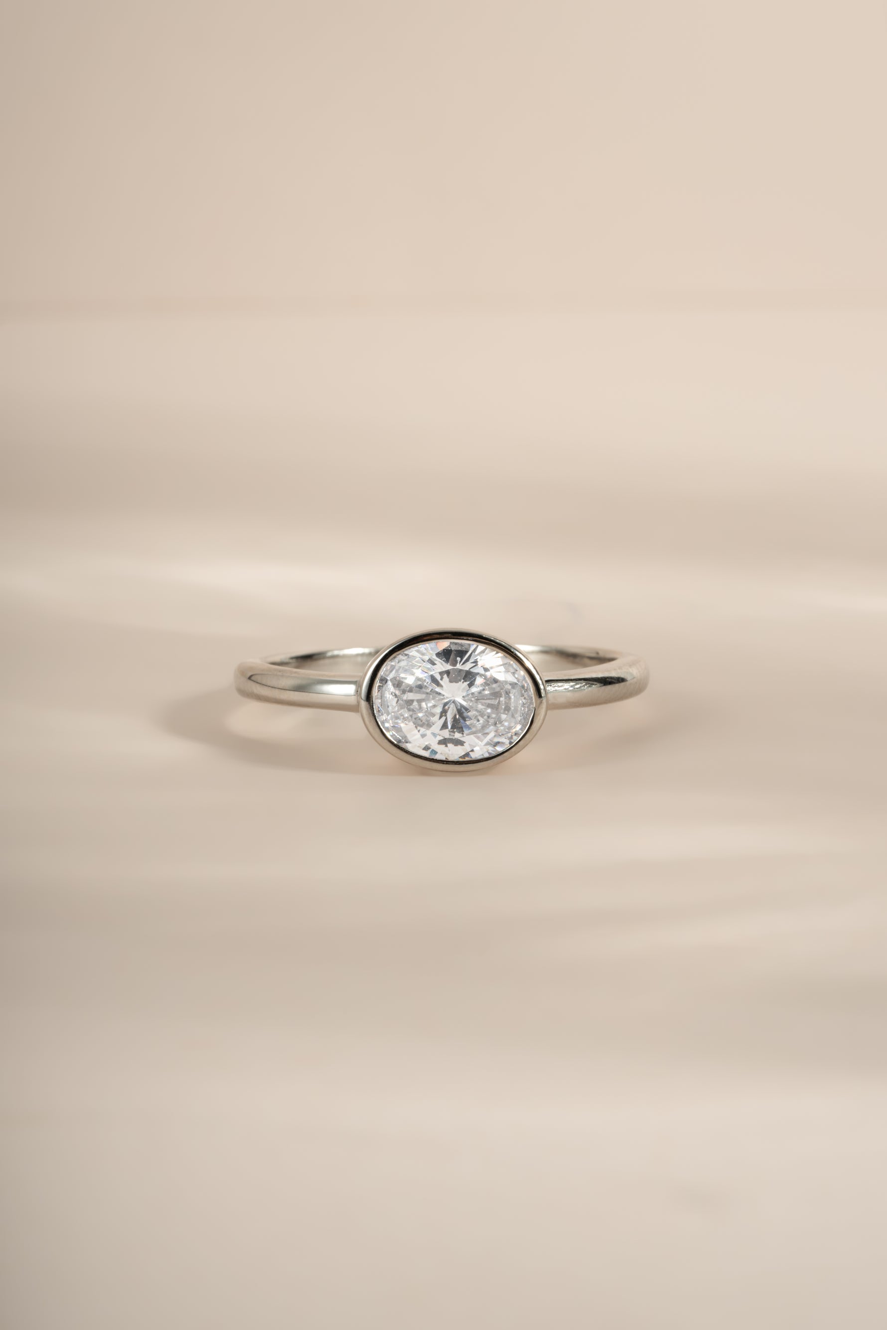 A silver ring with a bezel-set oval diamond gemstone, photographed on a soft beige background.