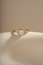 A gold ring with bezel oval solitaire diamond stone, displayed on a textured beige surface against a warm beige background.