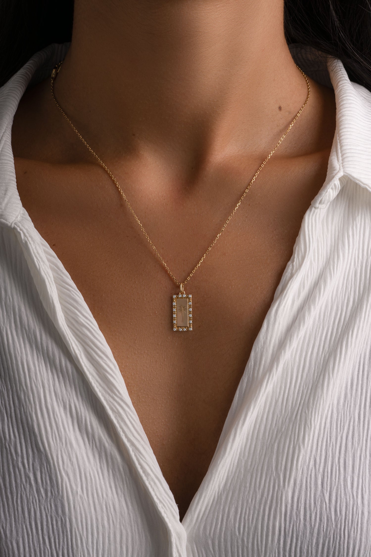 A close-up of a model wearing a delicate gold chain necklace with a small rectangular bezel-set pendant engraved with a palm tree, styled with a white textured blouse.