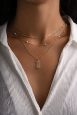 A close-up of a model wearing a layered gold necklace set, featuring a small rectangular pendant with a pavé setting and delicate pearl accents on the chains, styled with a white button-down shirt.