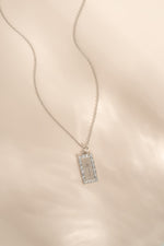 A silver-toned rectangular pendant necklace with a pavé-set border and a palm tree motif displayed on a soft beige fabric background.