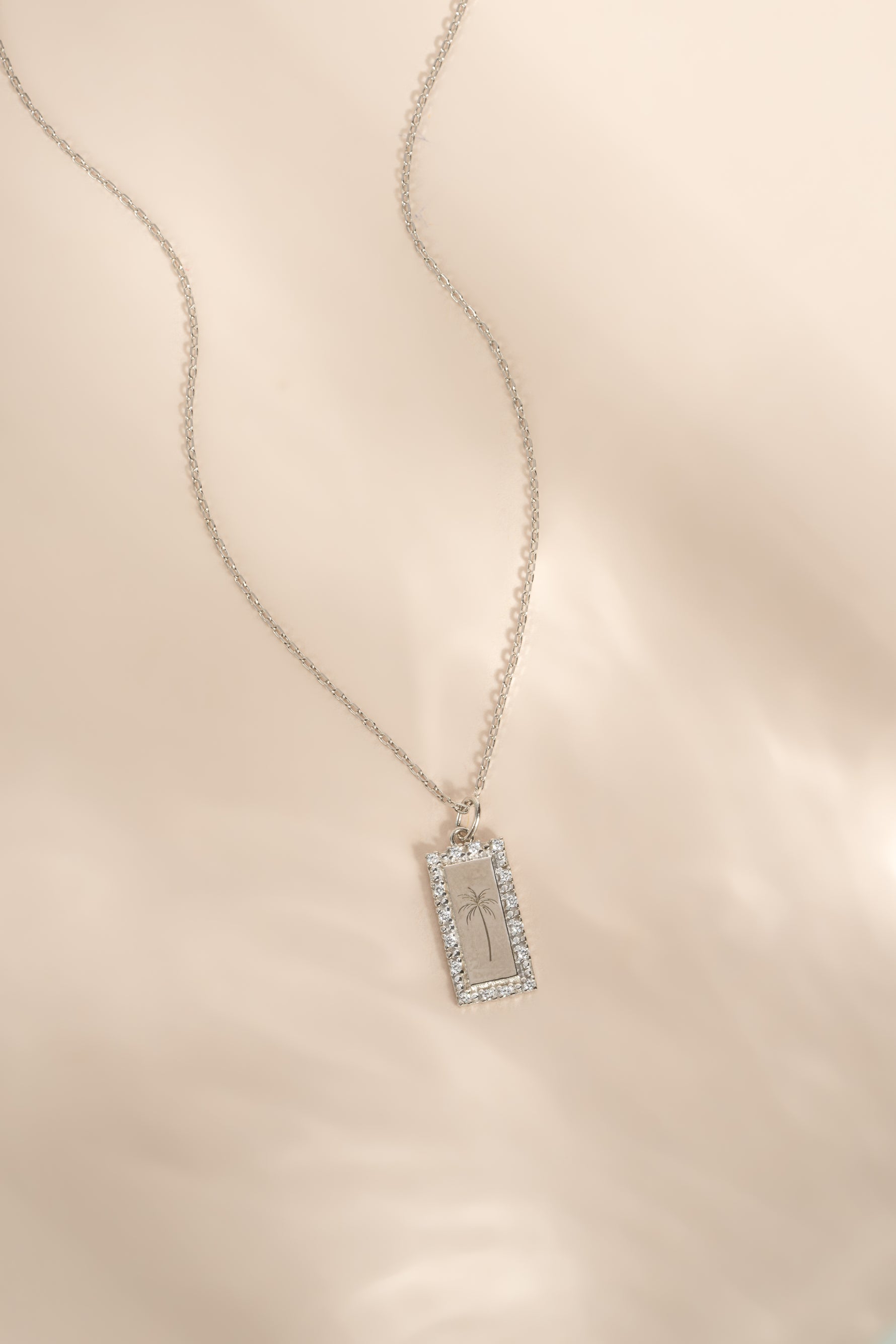 A silver-toned rectangular pendant necklace with a pavé-set border and a palm tree motif displayed on a soft beige fabric background.