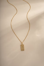 A gold chain necklace with a rectangular pendant featuring a palm tree and small diamond stones around the border, displayed on a warm beige background with soft shadows.