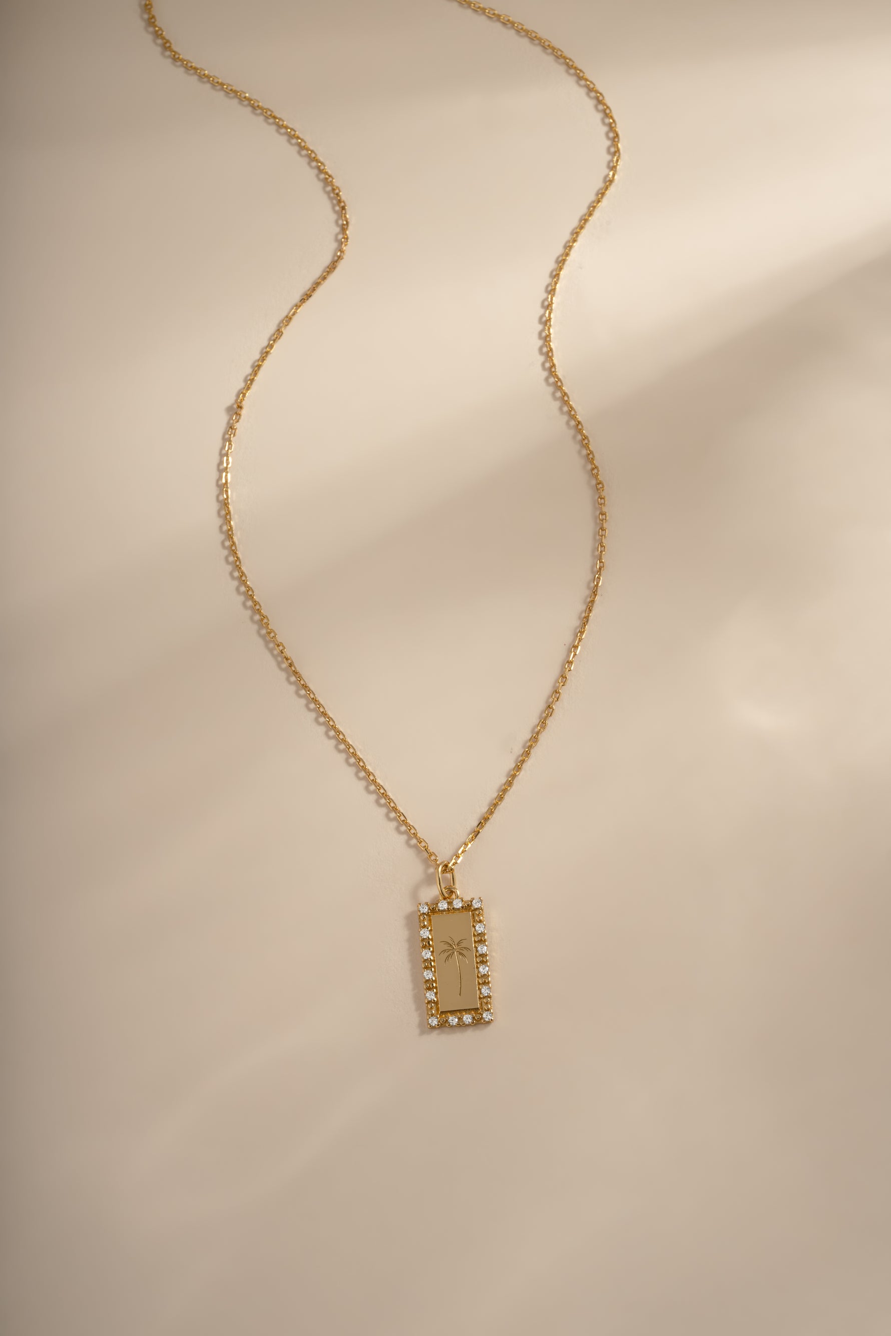 A gold chain necklace with a rectangular pendant featuring a palm tree and small diamond stones around the border, displayed on a warm beige background with soft shadows.