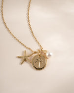 A gold-toned chain necklace featuring a palm tree pendant, a starfish charm, and a white pearl bead, displayed on a soft beige background.