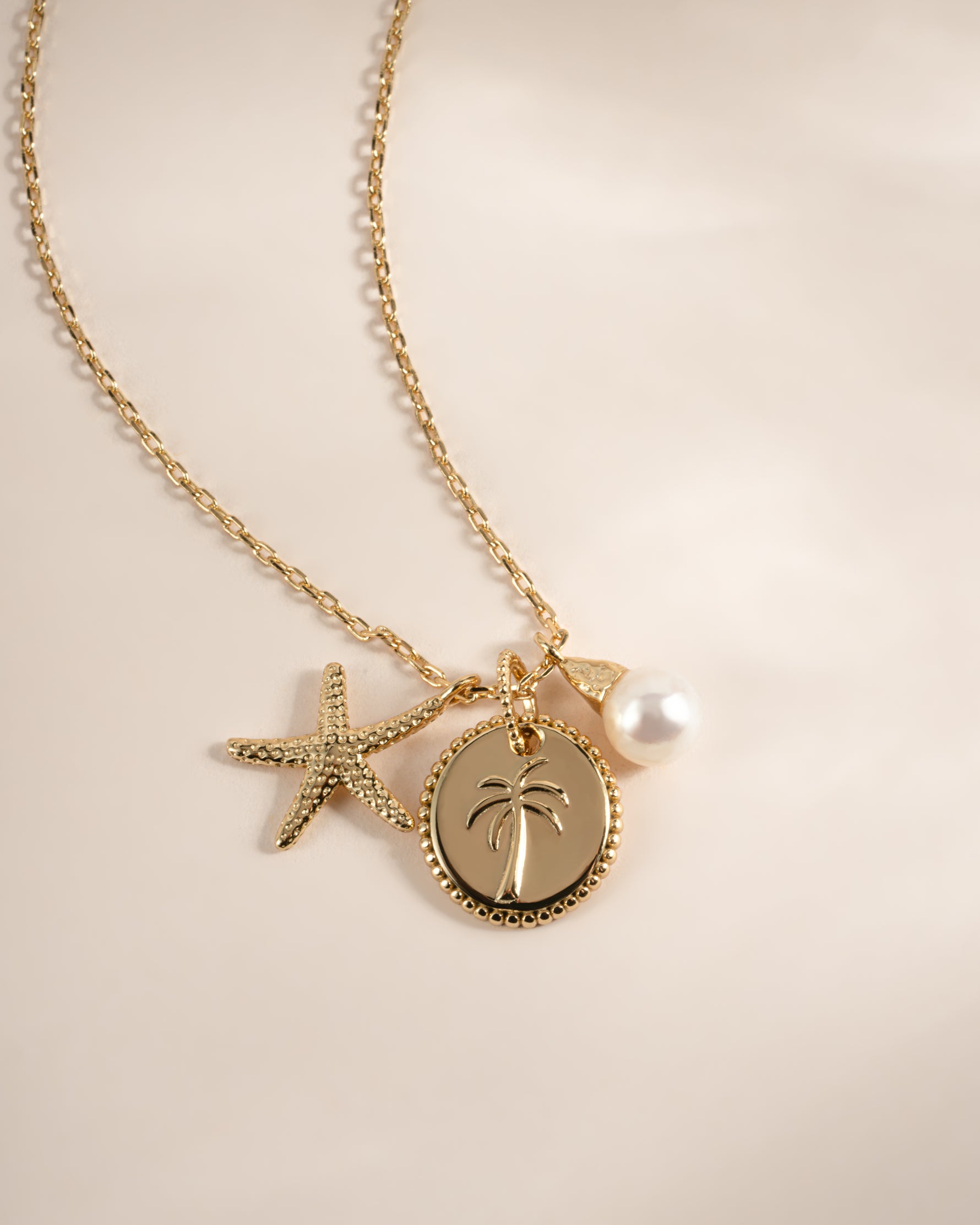 A gold-toned chain necklace featuring a palm tree pendant, a starfish charm, and a white pearl bead, displayed on a soft beige background.