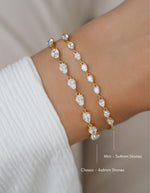 Two gold-toned bracelets with teardrop-shaped cz stones worn on a wrist, shown close up against a soft neutral background. Text labels indicate 'Mini - 5x4mm Stones' and 'Classic - 4x6mm Stones'.