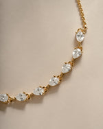 A close-up of a gold chain bracelet featuring a series of pear-cut clear gemstones in prong settings, arranged diagonally on a warm beige background.