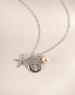 A silver-toned necklace with a delicate chain, featuring a starfish charm, a round palm tree pendant, and a small white pearl charm, displayed on a soft beige background.