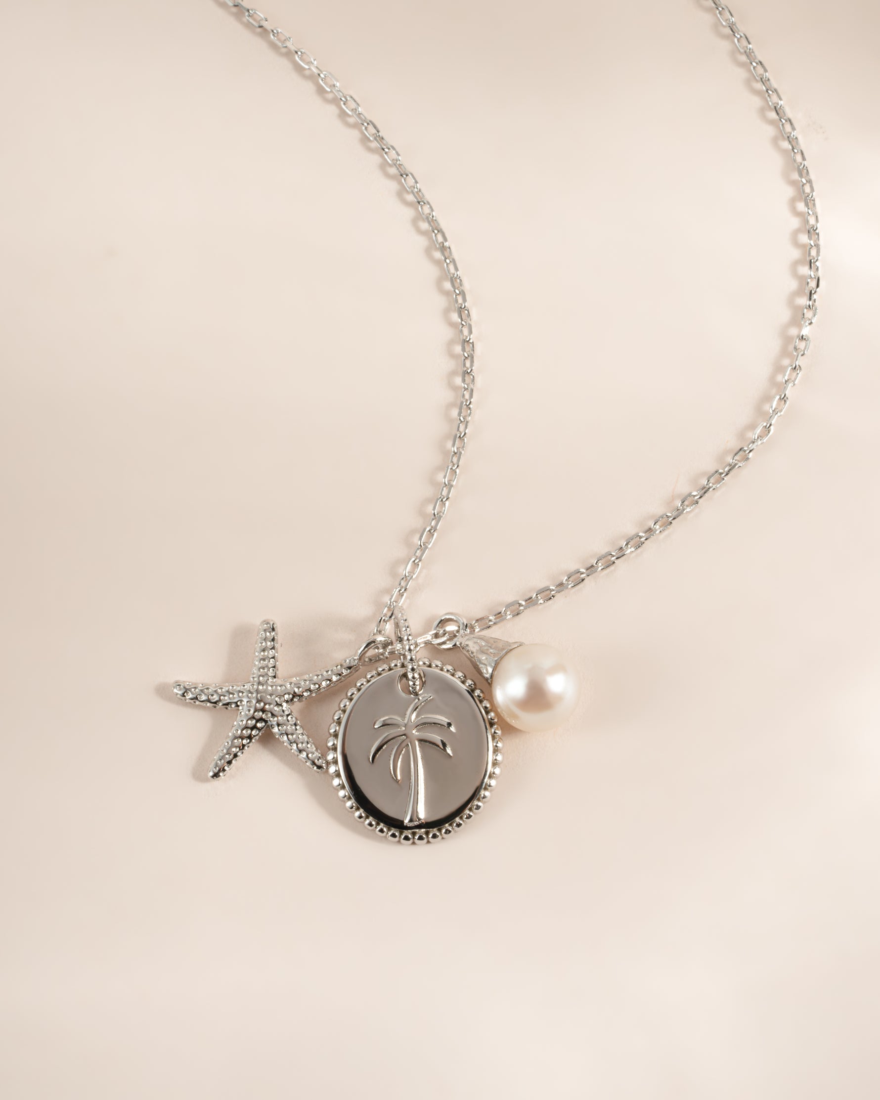 A silver-toned necklace with a delicate chain, featuring a starfish charm, a round palm tree pendant, and a small white pearl charm, displayed on a soft beige background.
