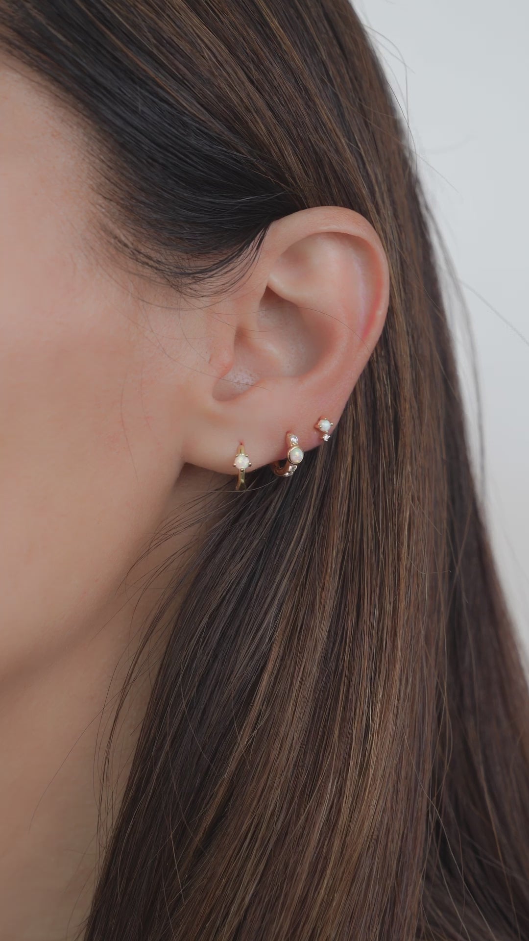 A closeup video of a model wearing two baby hoop earrings and one stud earrings. In the first piercing she is wearing huggie hoop earrings, in the second is another small opal earring with diamonds, and in the third is a tiny opal stud earring. 