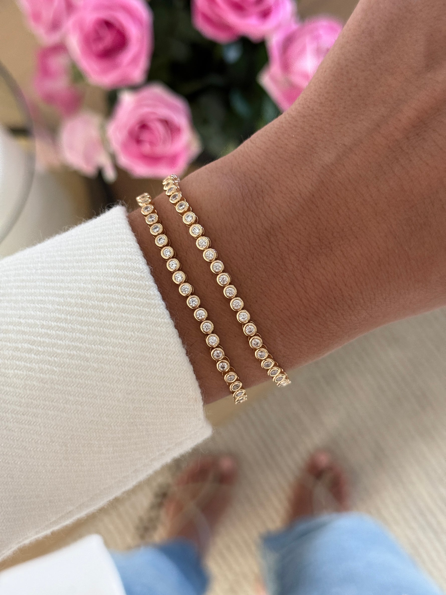 A close-up of a person's wrist wearing two delicate gold bracelets adorned with small, round, clear stones. The person is wearing a white ribbed knit sweater, and a blurred background of pink roses is visible.