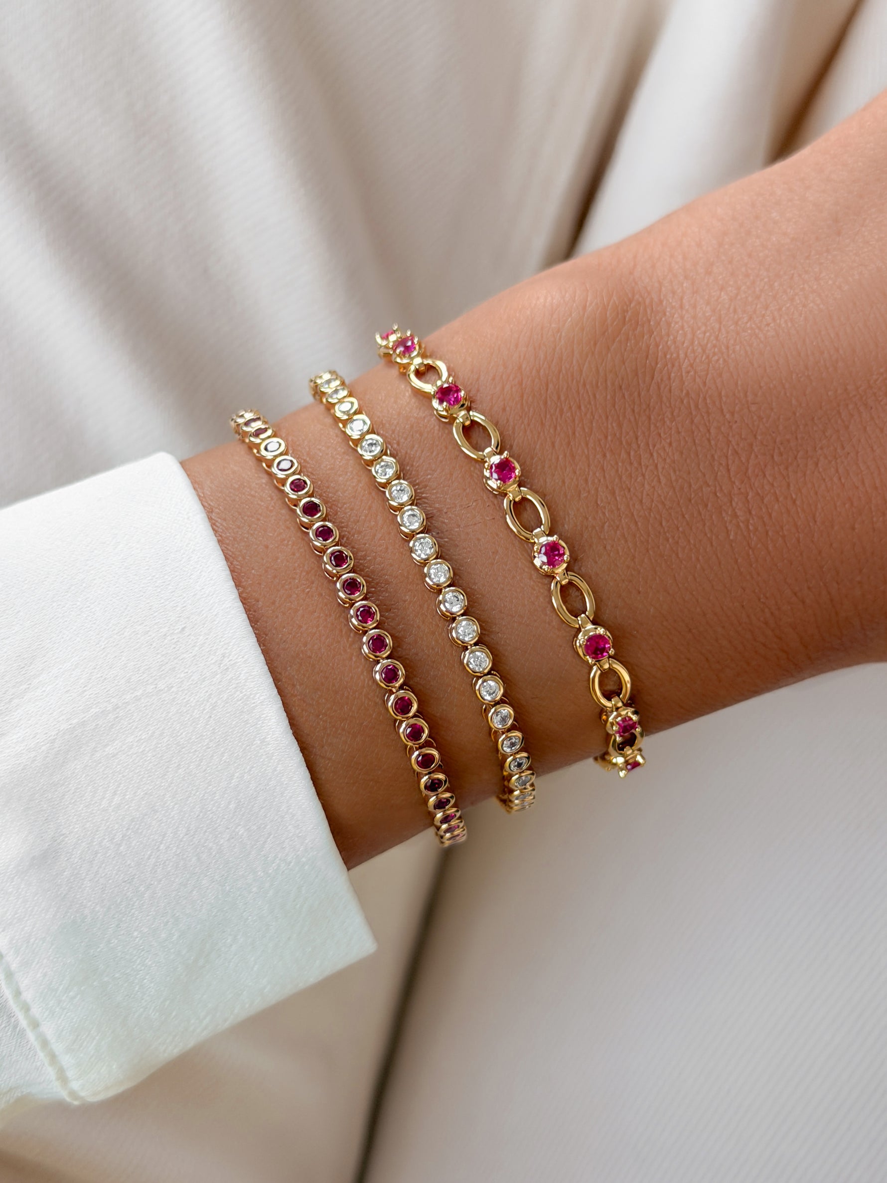 Three delicate gold bracelets with pink gemstones and clear stones, worn stacked on a person's wrist against a soft cream-colored background.