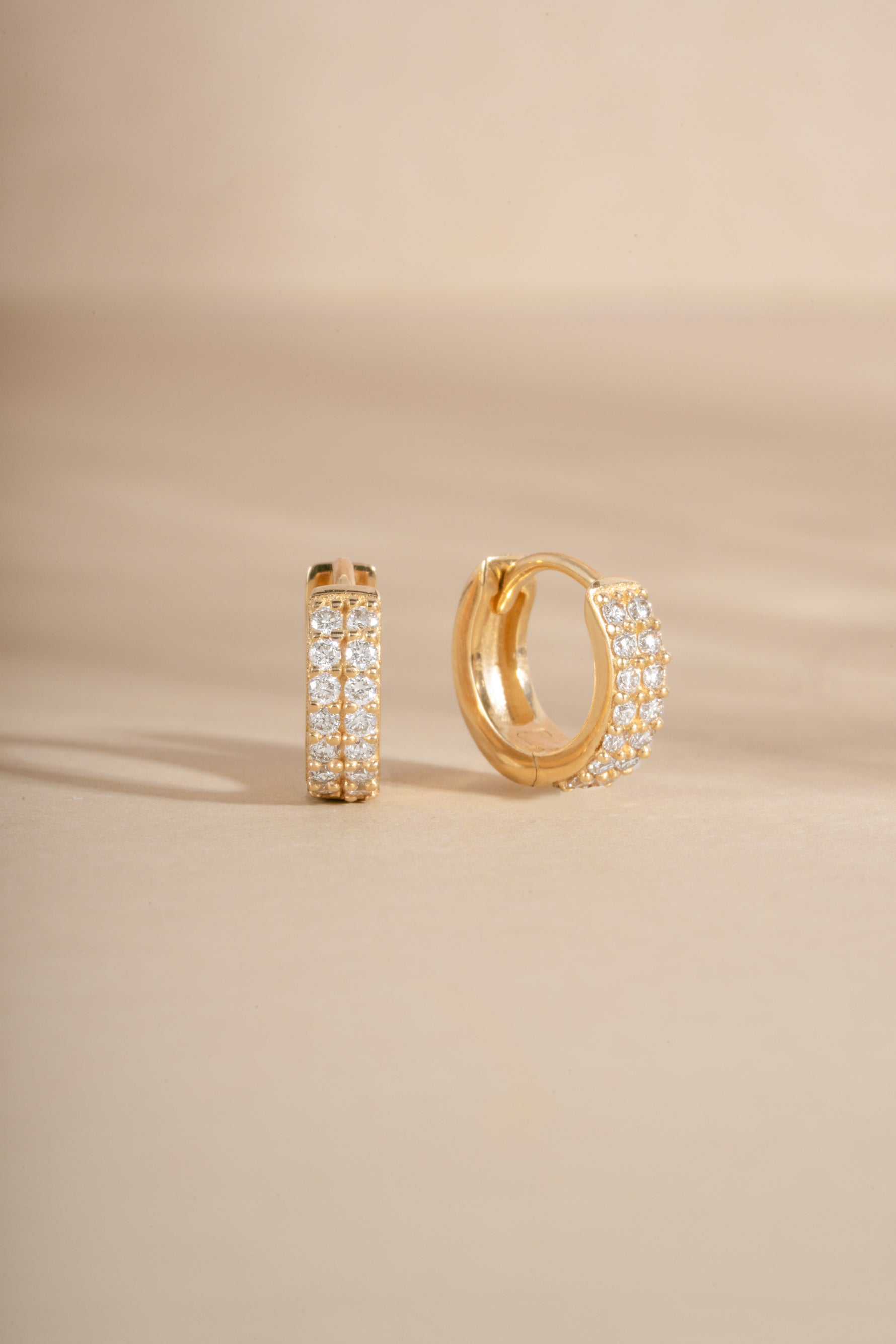 A pair of gold huggie hoop earrings adorned with pavé-set clear stones, displayed on a warm beige background.