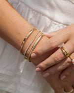 Close-up of a hand wearing multiple gold stacking bracelets and rings against a blurred white background, bracelets are featuring a gold diamond bangle bracelet, a gold tennis bracelet, and a bezel set diamond bangle