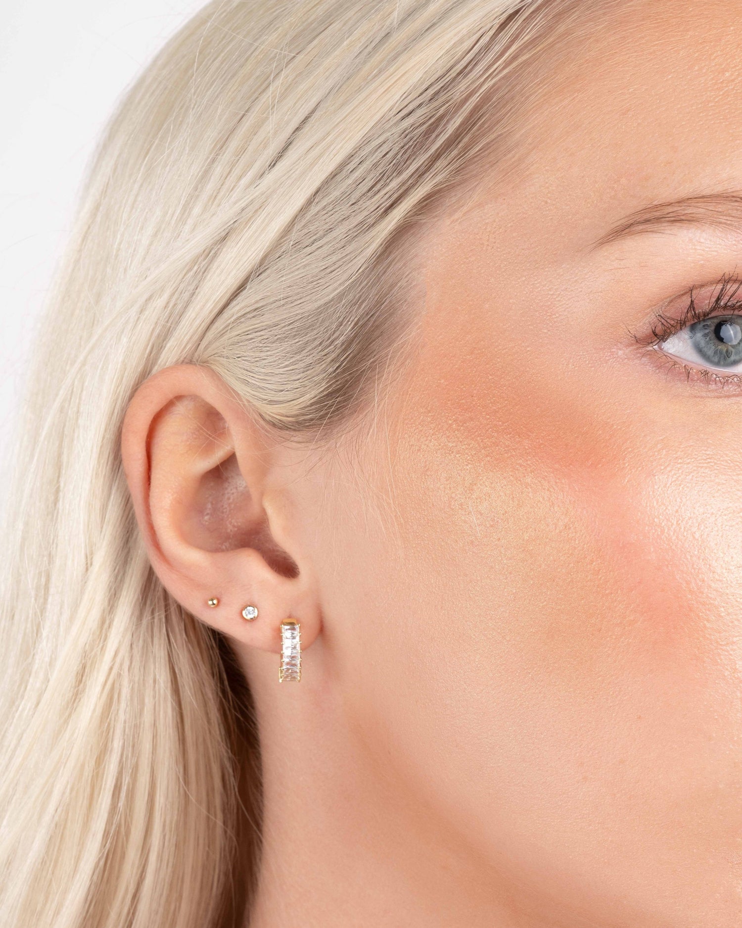 Model wearing gold baguette huggie hoops, a diamond stud earring, and a ball stud earring