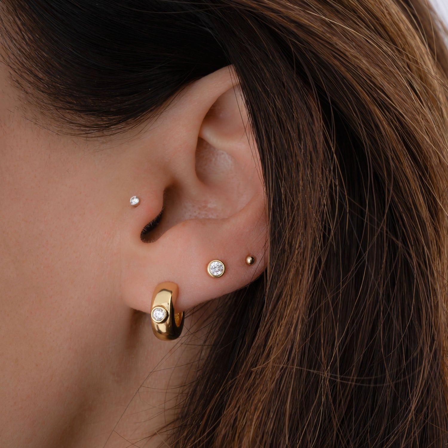 Model wearing chunky gold hoops, a diamond bezel stud earring, a ball stud earring in gold, and a solitaire stud in the tragus piercing. 