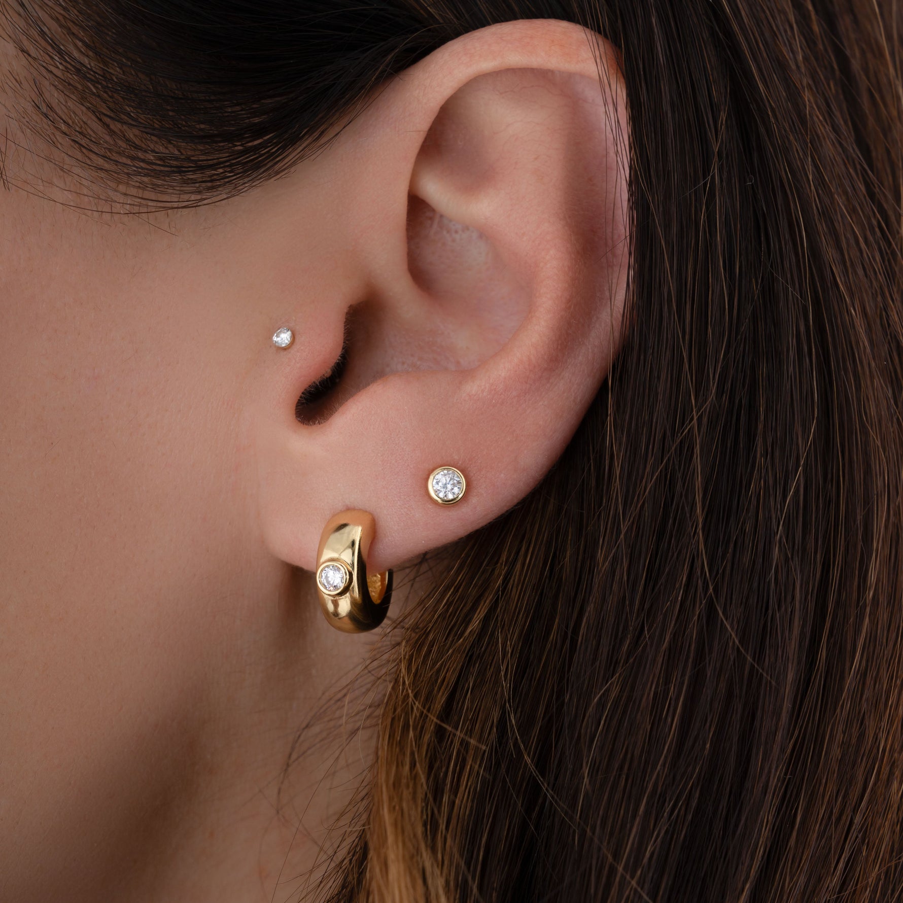 Model wearing chunky huggie hoop earrings plated in 14K gold, a pair of diamond bezel stud earrings, and a solitaire stud in the tragus piercing