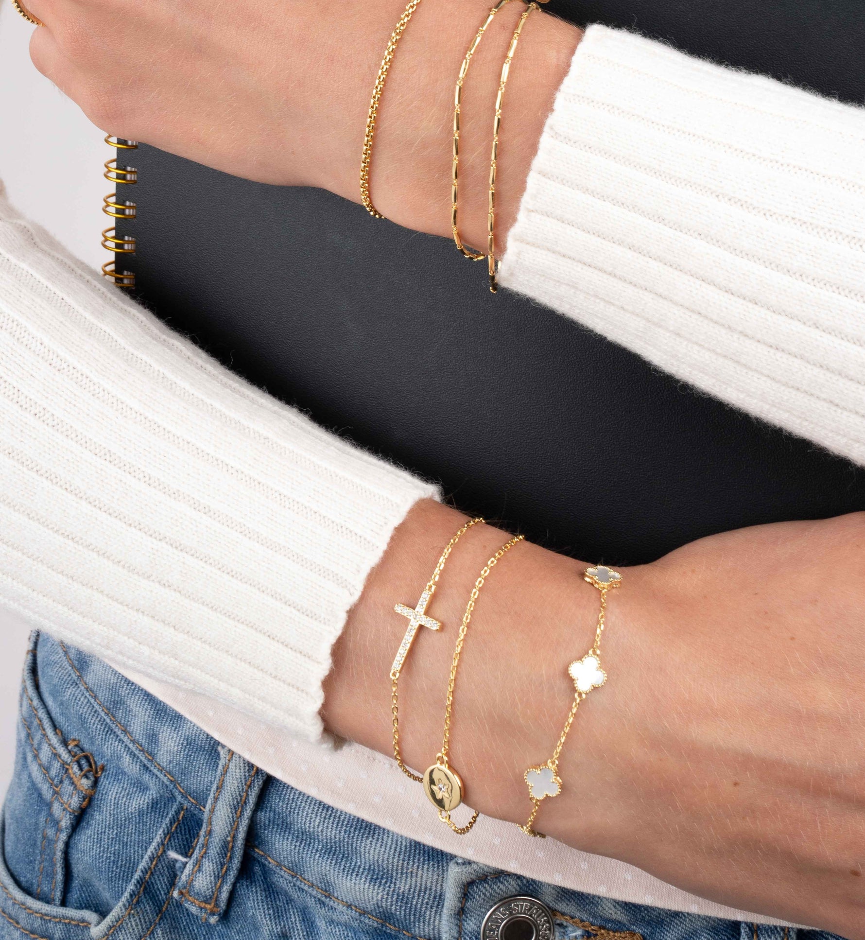 Model wearing gold bracelet stacks on each wrist while holding a spiral notebook, featuring a gold pearl bracelet, a gold chain bracelet and a gold cross pendant adjustable bracelet