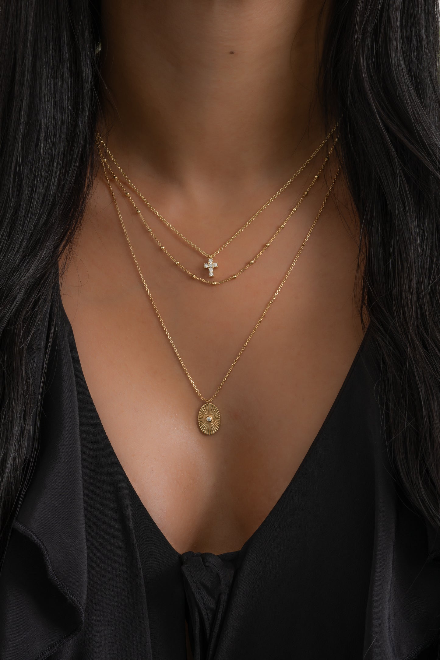 model wearing gold necklace stack including an oval medallion pendant necklace, a beaded chain necklace, and a small cross pendant necklace