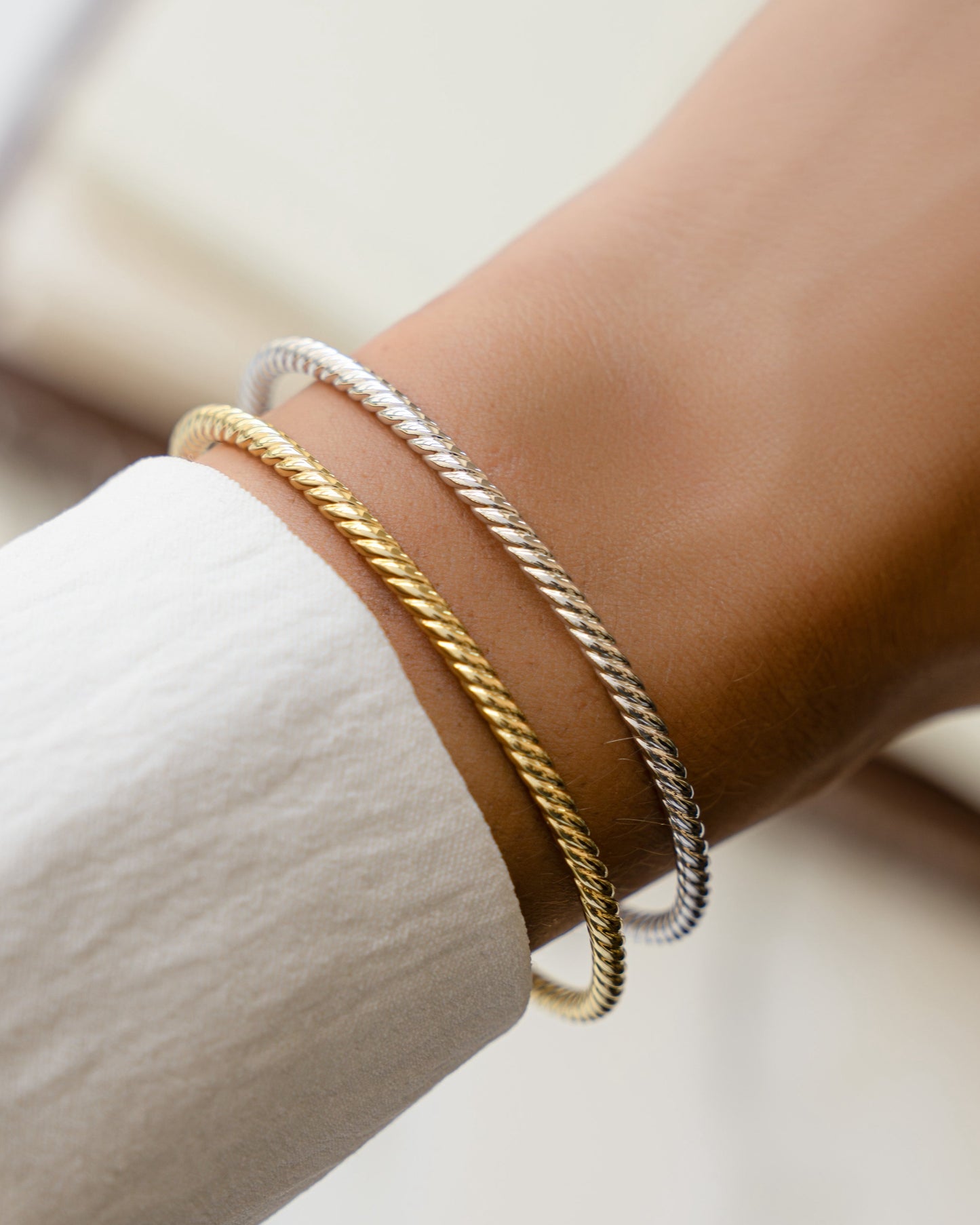 closeup image of model wearing two bangle bracelets with a twisted design in silver and in gold