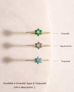 Three gold floral rings with gemstones labeled 'Emerald', 'Aquamarine', and 'Turquoise' on a beige background.